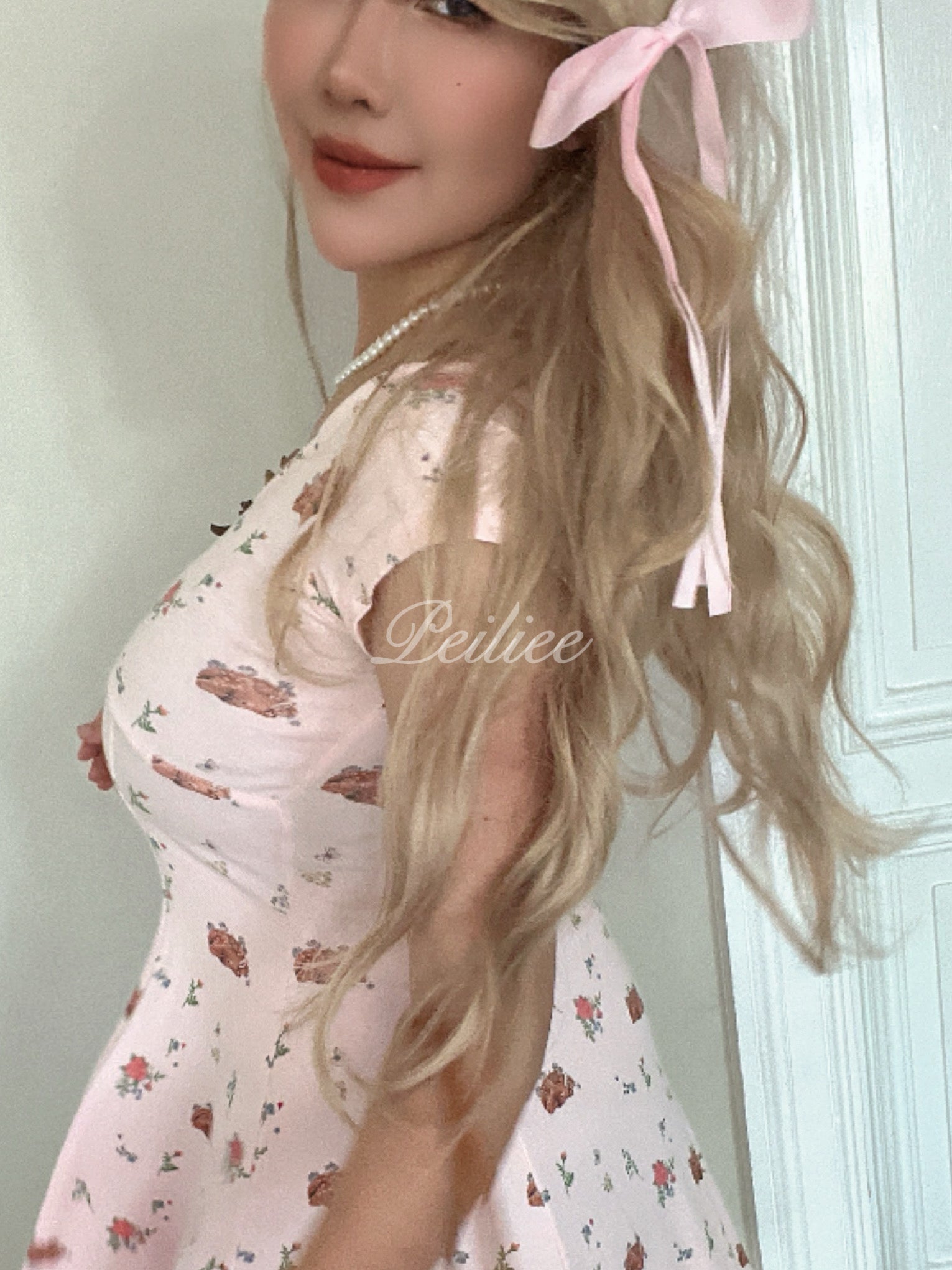 Woman with long blonde hair wearing a pink floral dress, standing against a light-colored wall.