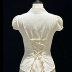 An off-white short-sleeve blouse with a lace-up corset-style back, displayed on a cream mannequin against a black background.
