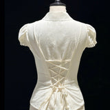 An off-white short-sleeve blouse with a lace-up corset-style back, displayed on a cream mannequin against a black background.