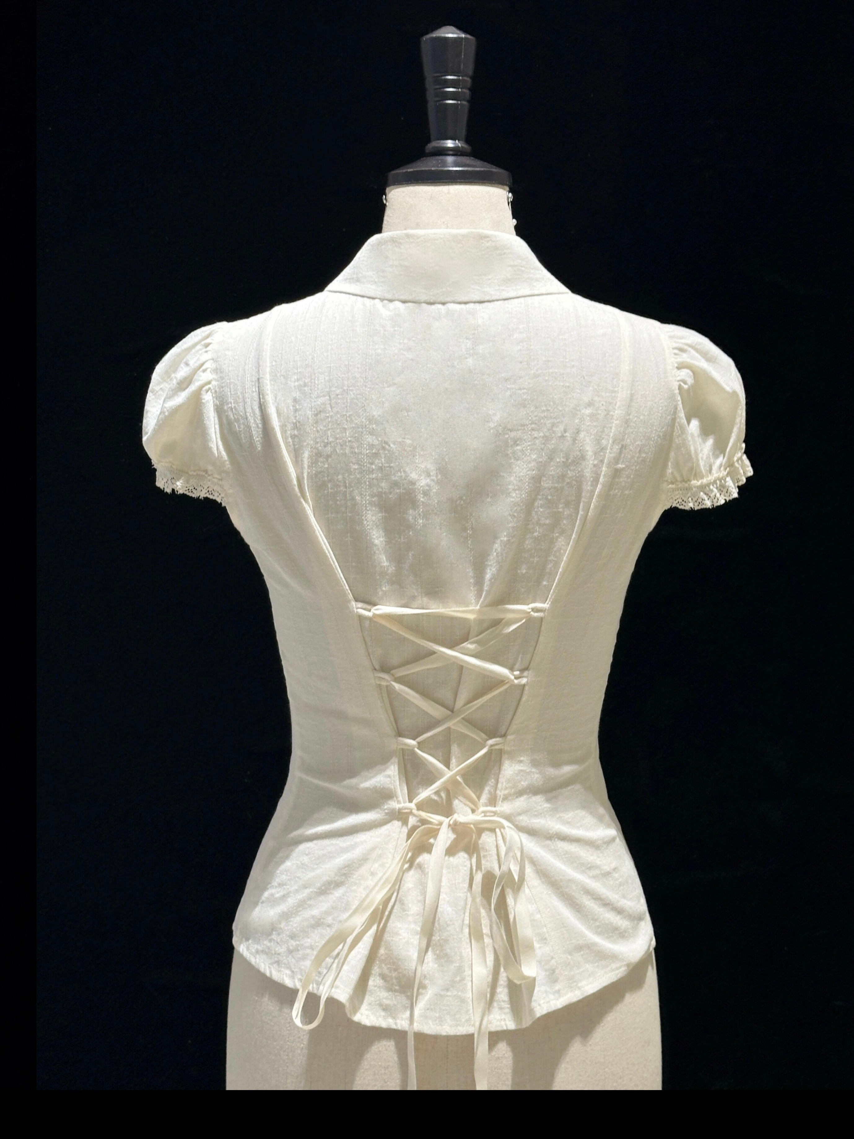An off-white short-sleeve blouse with a lace-up corset-style back, displayed on a cream mannequin against a black background.