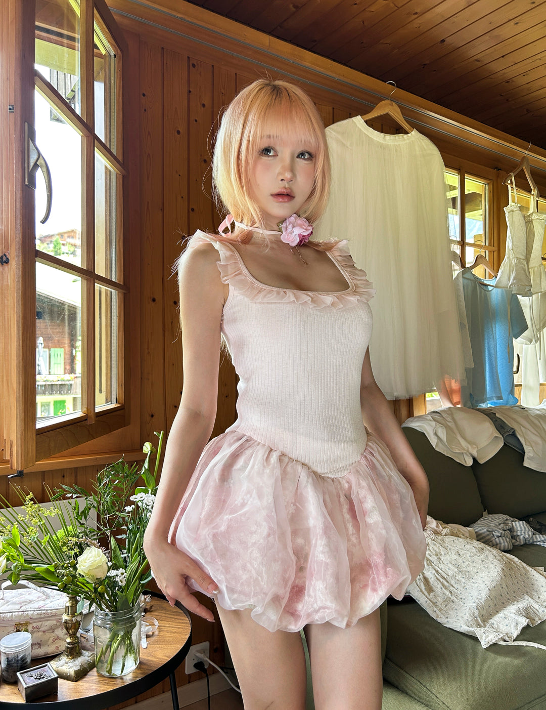 Teenager girl in pink hair wearing a pink dress standing in a room with wooden walls and a window.